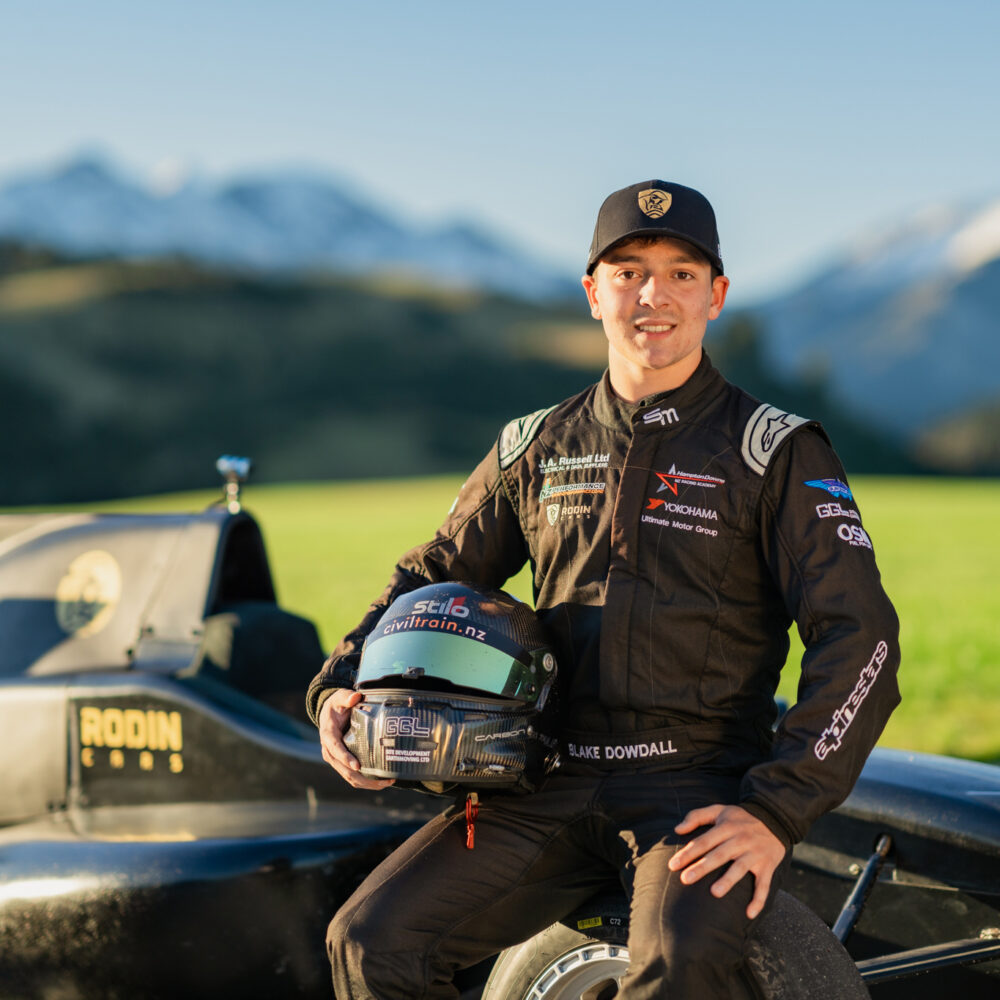 Blake Dowdall sits on edge of Formula Ford car with helmet by his side smiling at the camera. Mt Lyford is in the background with snow caps.