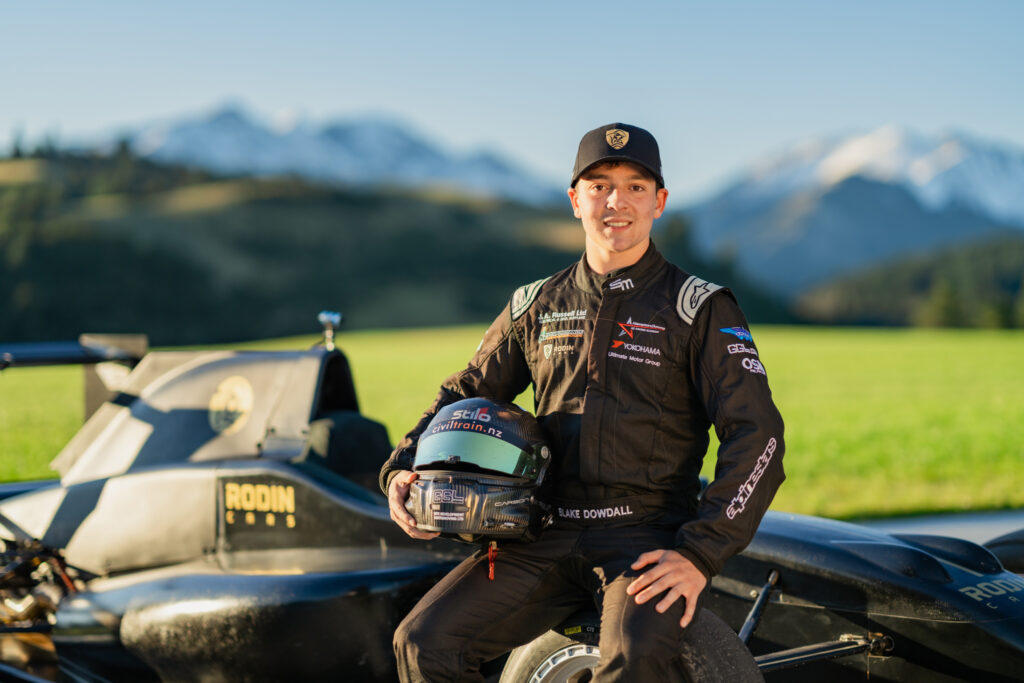 Blake Dowdall sits on edge of Formula Ford car with helmet by his side smiling at the camera. Mt Lyford is in the background with snow caps.