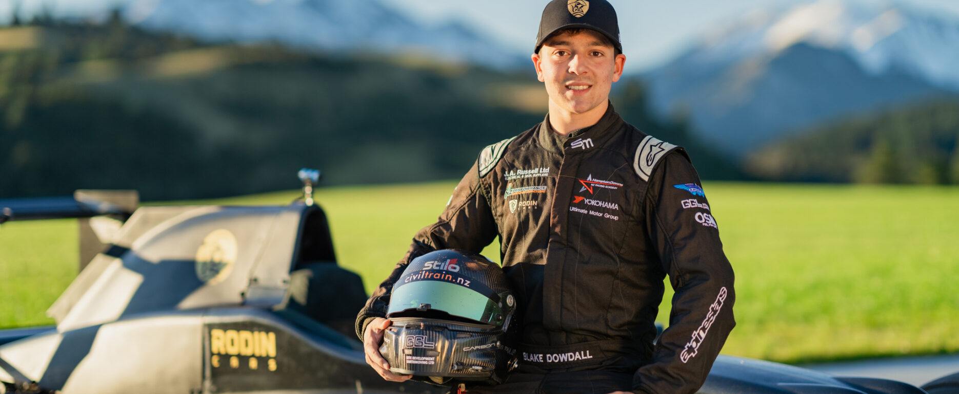 Blake Dowdall sits on edge of Formula Ford car with helmet by his side smiling at the camera. Mt Lyford is in the background with snow caps.